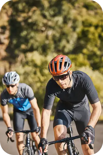 Deux cyclistes portant un casque et des lunettes de soleil roulent côte à côte sur une route ensoleillée, le visage concentré. Feuilleillage vert flou en arrière-plan.
